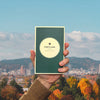 Hand holding a book titled 'Portland' against a cityscape with autumn trees and mountains.
